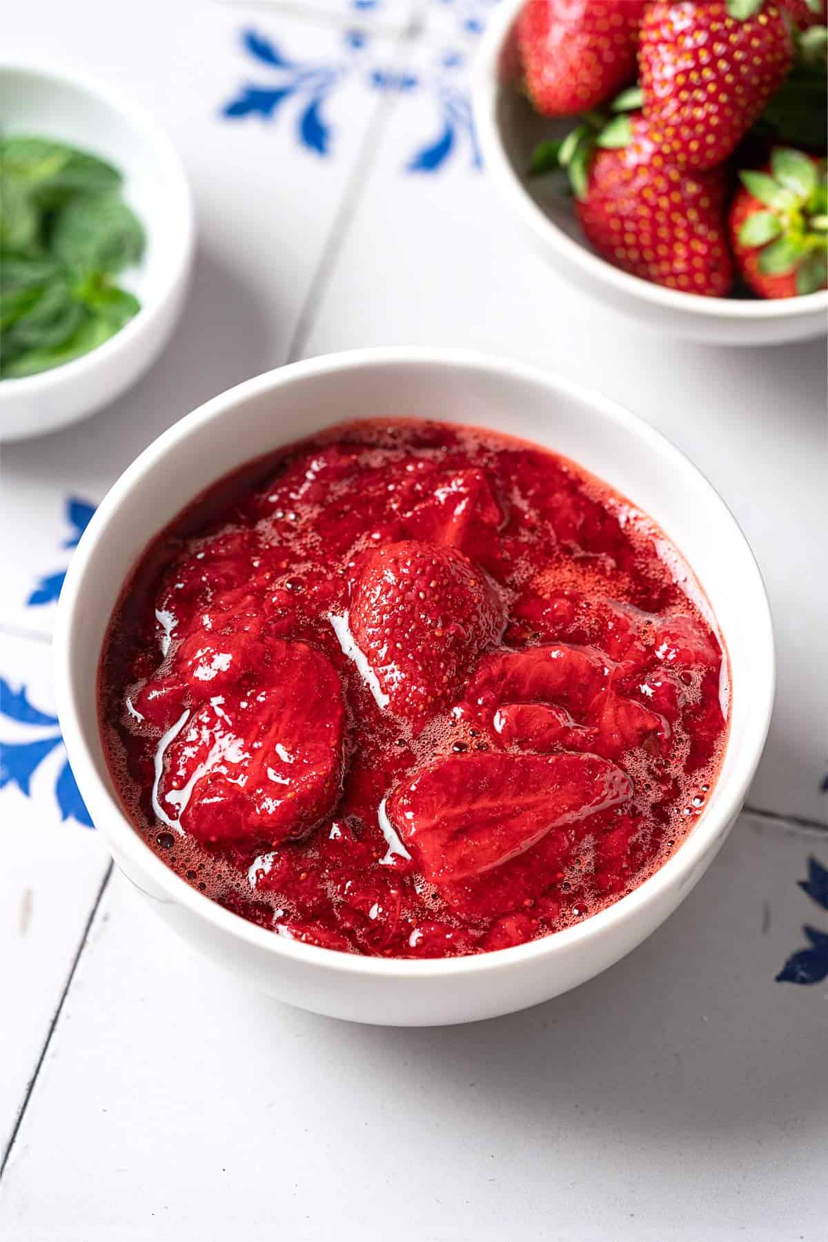 strawberry compote in a bowl next to a bowl of strawberries and a bowl of mint.