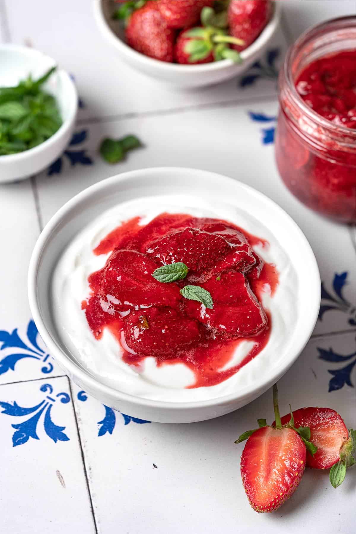 yogurt topped with strawberry compote in a bowl next to 2 strawberry halves, a jar of strawberry compote, a bowl of strawberries and a bowl of mint.