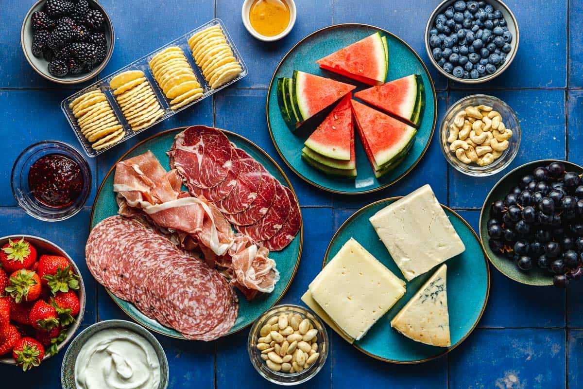 ingredients for a fruit charcuterie board including blackberries, watermelon, blueberries, grapes, strawberries, crackers, cheeses, assorted Italian meats, almonds, cashews, greek yogurt and honey.