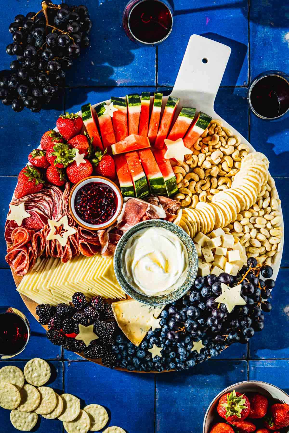 overhead photo of a complete fruit charcuterie board surrounded by 3 glasses of wine, grapes, a bowl of strawberries, and a group of crackers.