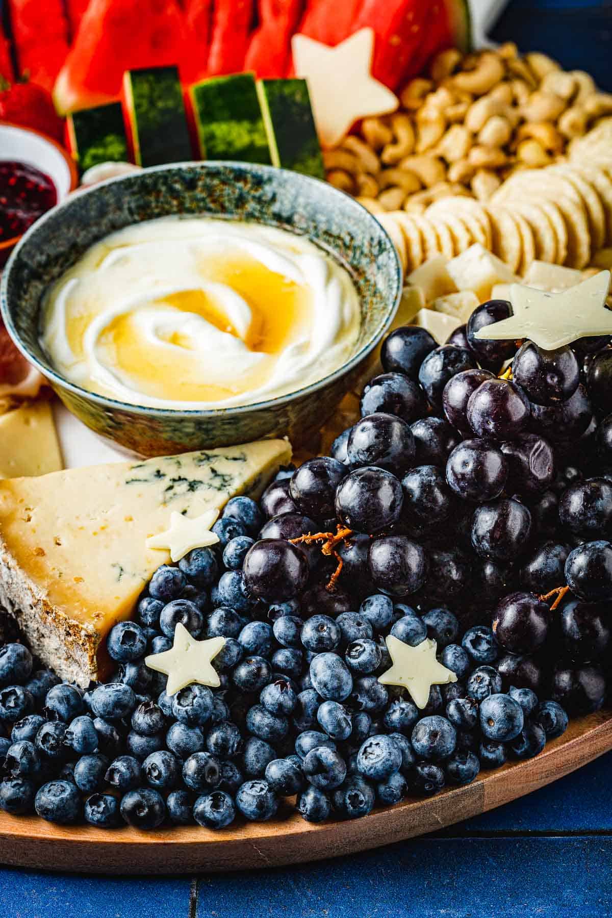 close up of a complete fruit charcuterie board highlighting cheese, blueberries, nuts, crackers, watermelon and bowl of greek yogurt topped with honey.