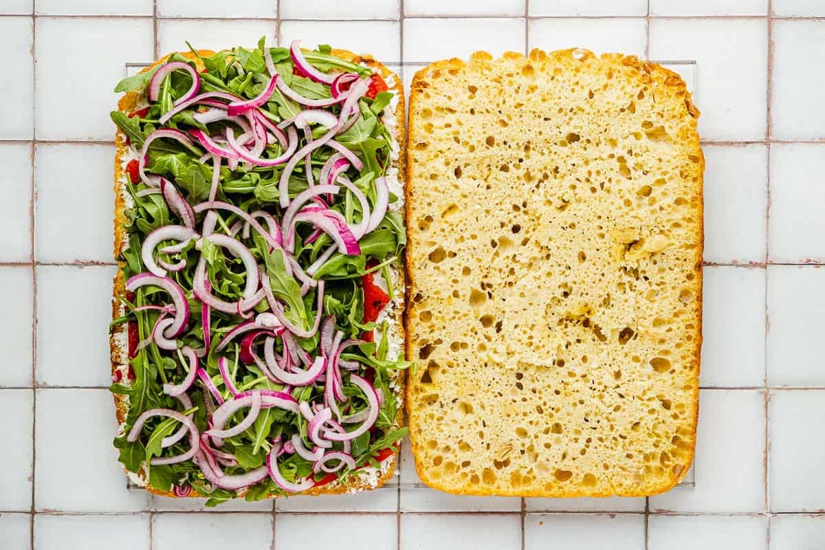 a focaccia loaf sliced in half with cheese, roasted red peppers, arugula and sliced red onions on one side.