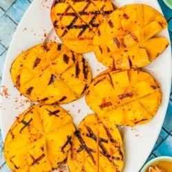 six grilled mango halves on a serving platter next to bowls of kosher salt, aleppo pepper, and honey.