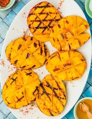 six grilled mango halves on a serving platter next to bowls of kosher salt, aleppo pepper, and honey.