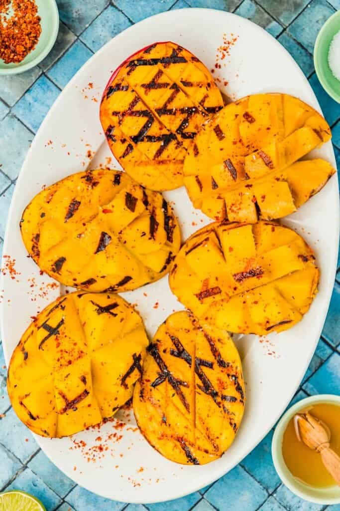 six grilled mango halves on a serving platter next to bowls of kosher salt, aleppo pepper, and honey.