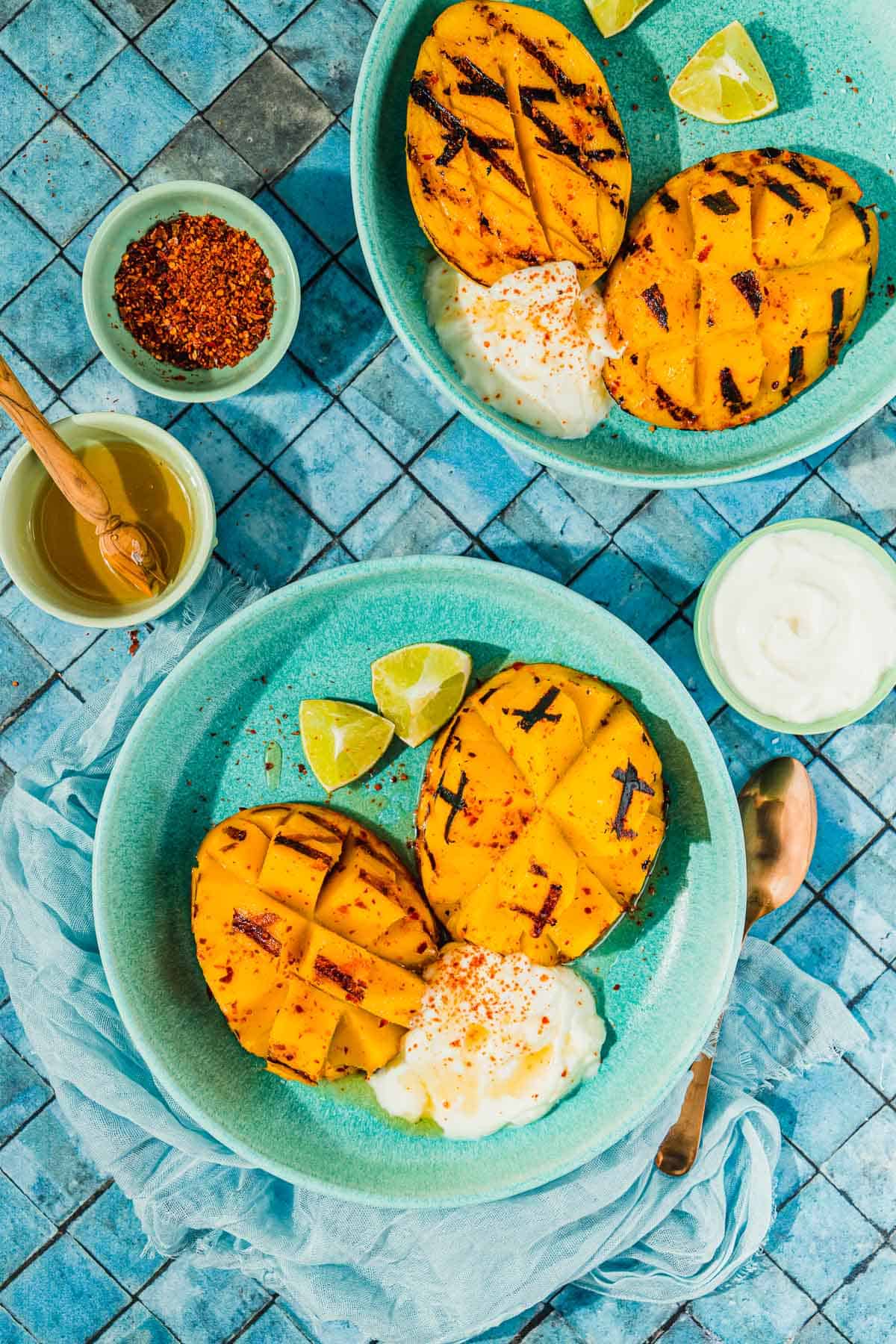 two bowls with two grilled mango halves, a dollop of whipped cream and 2 lime wedges in each next to a bowl of honey, a bowl of whipped cream, and a bowl of aleppo pepper.