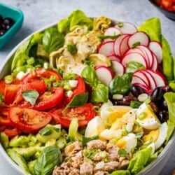 An entire nicoise salad in a serving bowl next to a bowl of artichoke hearts and a bowl of sliced tomatoes.