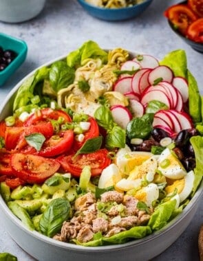 An entire nicoise salad in a serving bowl next to a bowl of artichoke hearts and a bowl of sliced tomatoes.