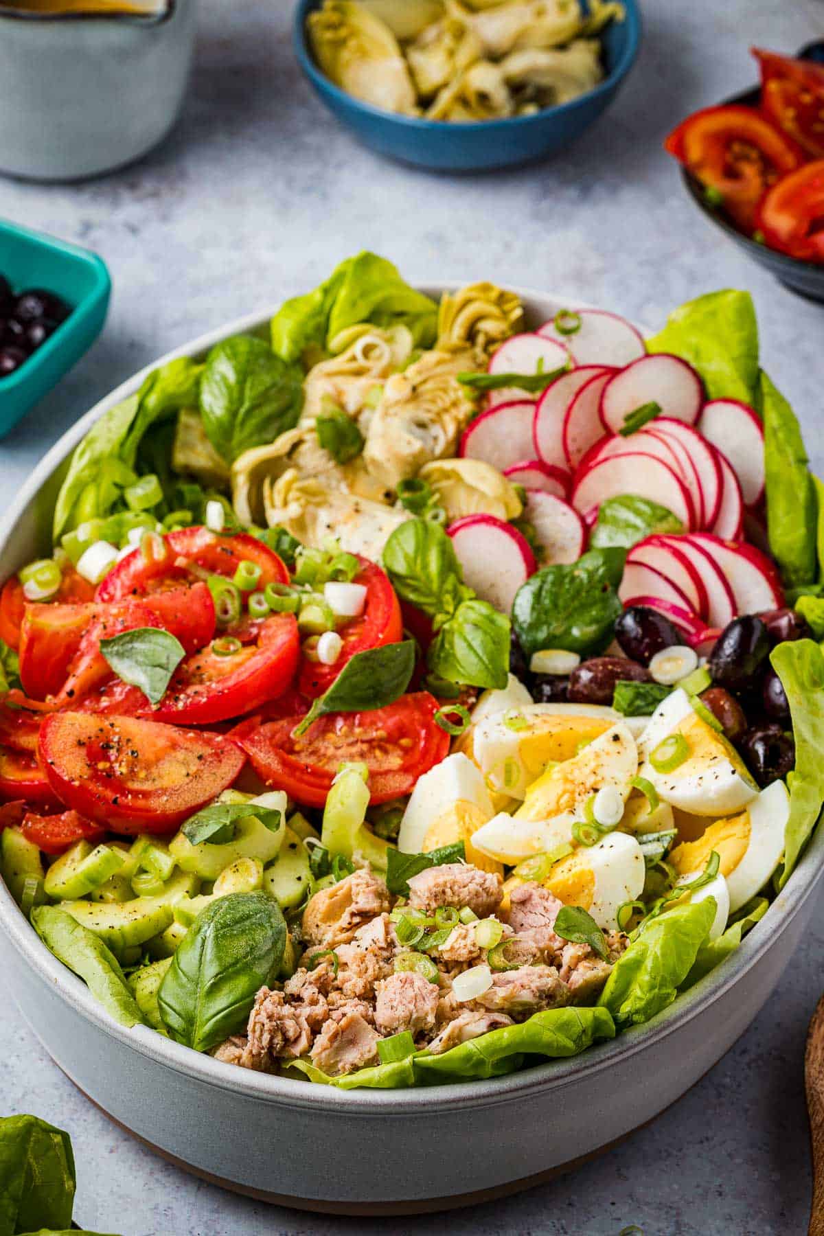 An entire nicoise salad in a serving bowl next to a bowl of artichoke hearts and a bowl of sliced tomatoes.