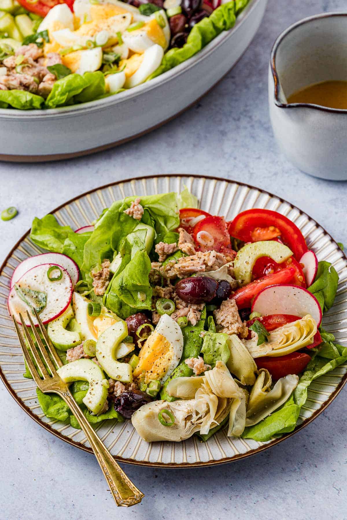 a serving of nicoise salad on a plate with a fork, next to a cup of salad dressing and an entire nicoise salad in a serving bowl.