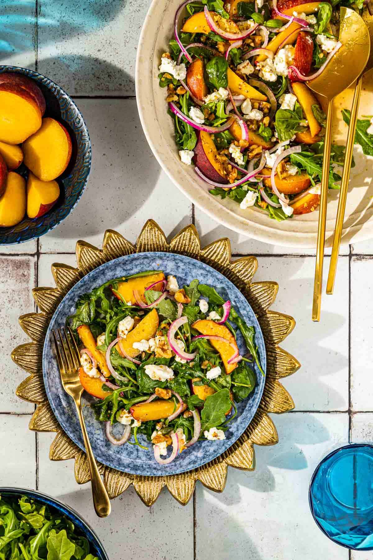 overhead photo of a peach salad on a plate with a fork in front of an entire peach salad in a serving bowl with gold serving utensils, a bowl of peaches, a bowl of arugula and a glass of water.