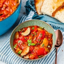 ratatouille in a bowl next to a spoon, a pot of ratatouille, and slices of toasted bread.