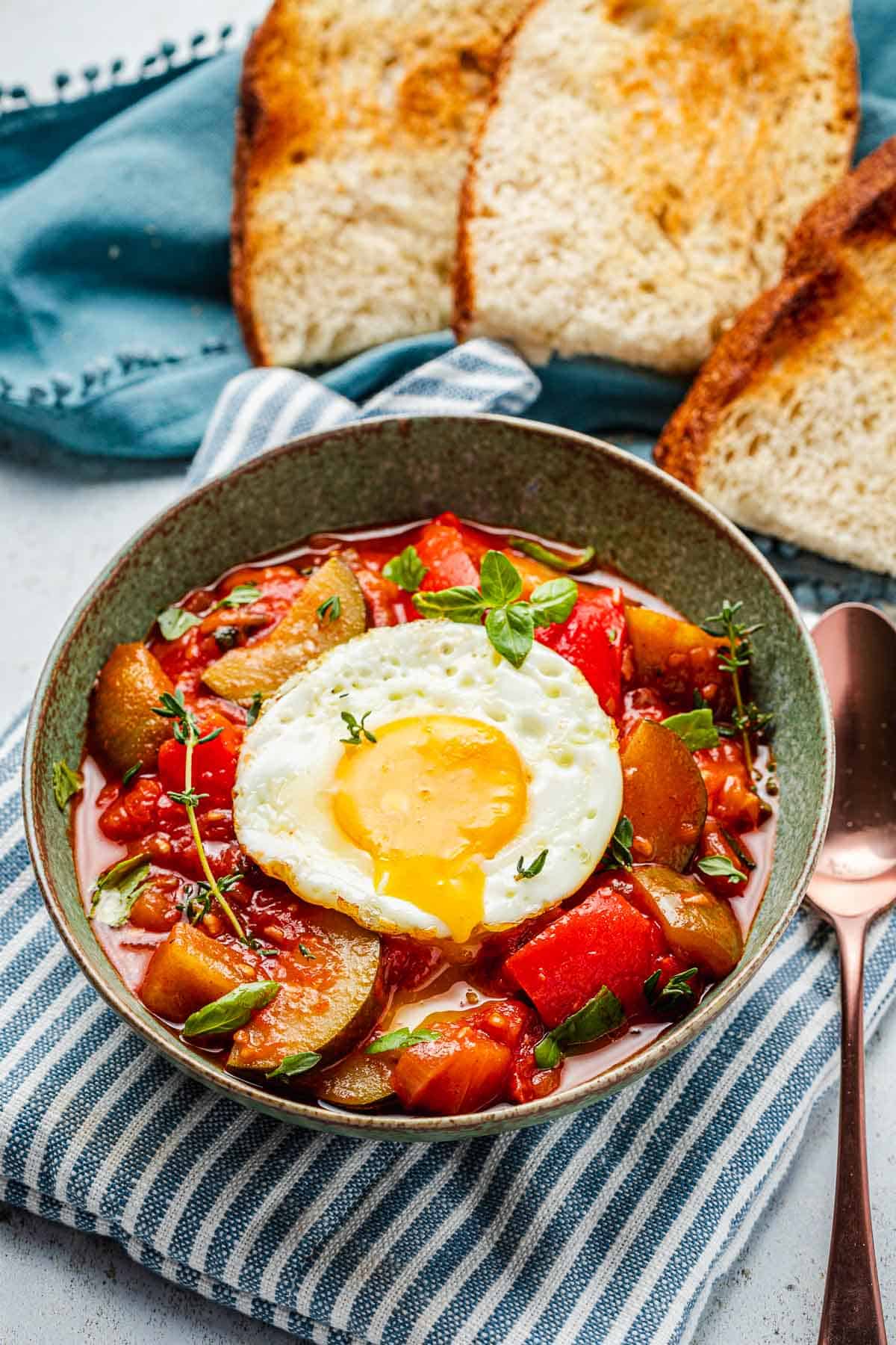 ratatouille topped with a fried egg in a bowl next to a spoon, and slices of toasted bread.