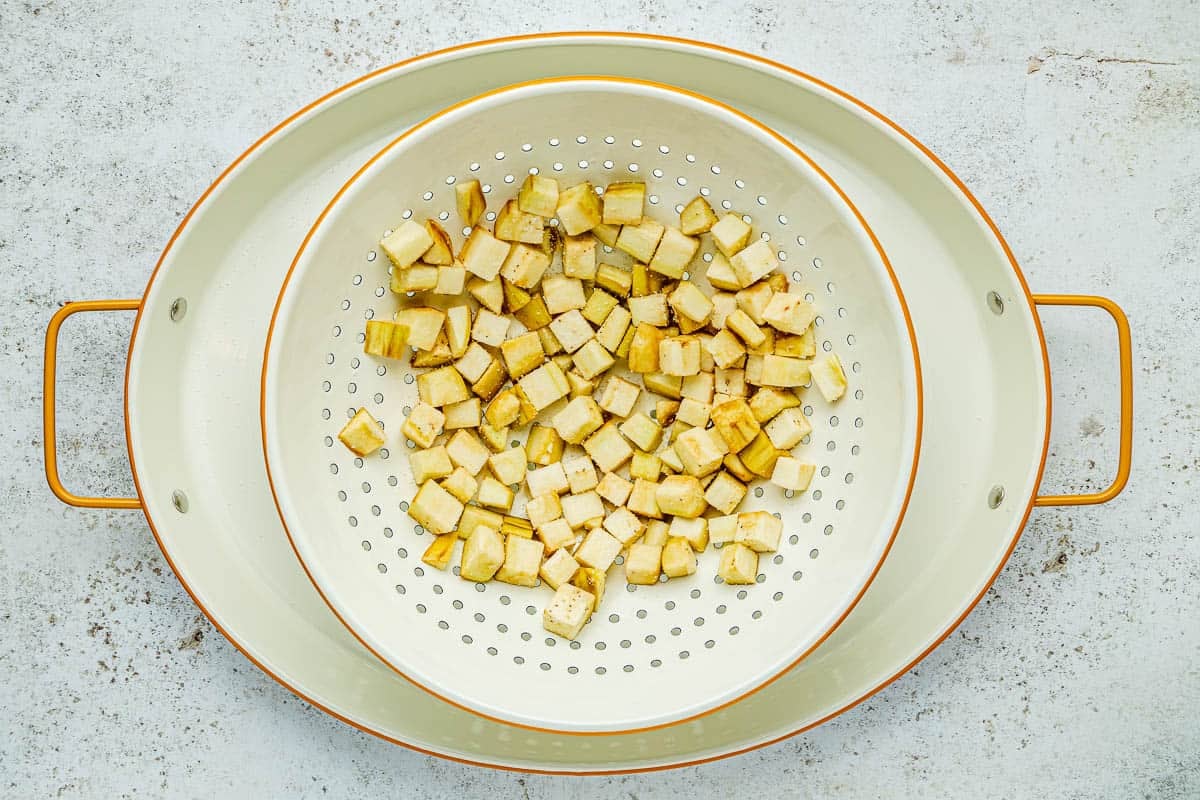 cubed and salted eggplant draining in a colander set inside a baking pan.