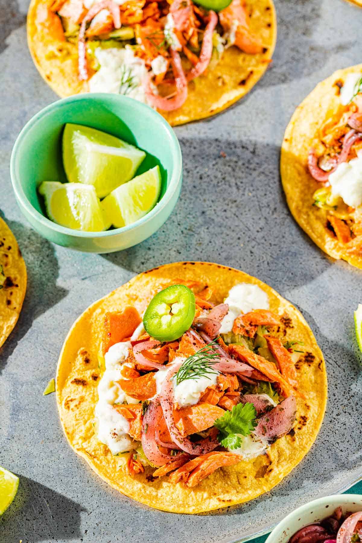 three open salmon tacos on corn tortillas topped with tzatziki, pickled onions and sliced jalapenos next to a bowl with 3 lime wedges.