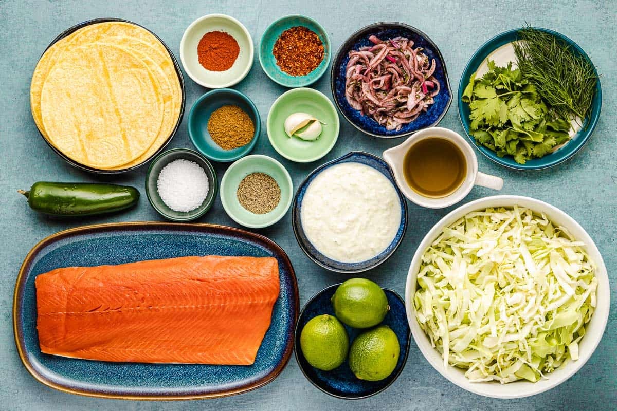 ingredients for salmon tacos including salmon, corn tortillas, salt, pepper, garlic, cumin, paprika, aleppo pepper, olive oil, limes, tzatziki sauce, pickled onions, jalapenos, cabbage, dill and cilantro.