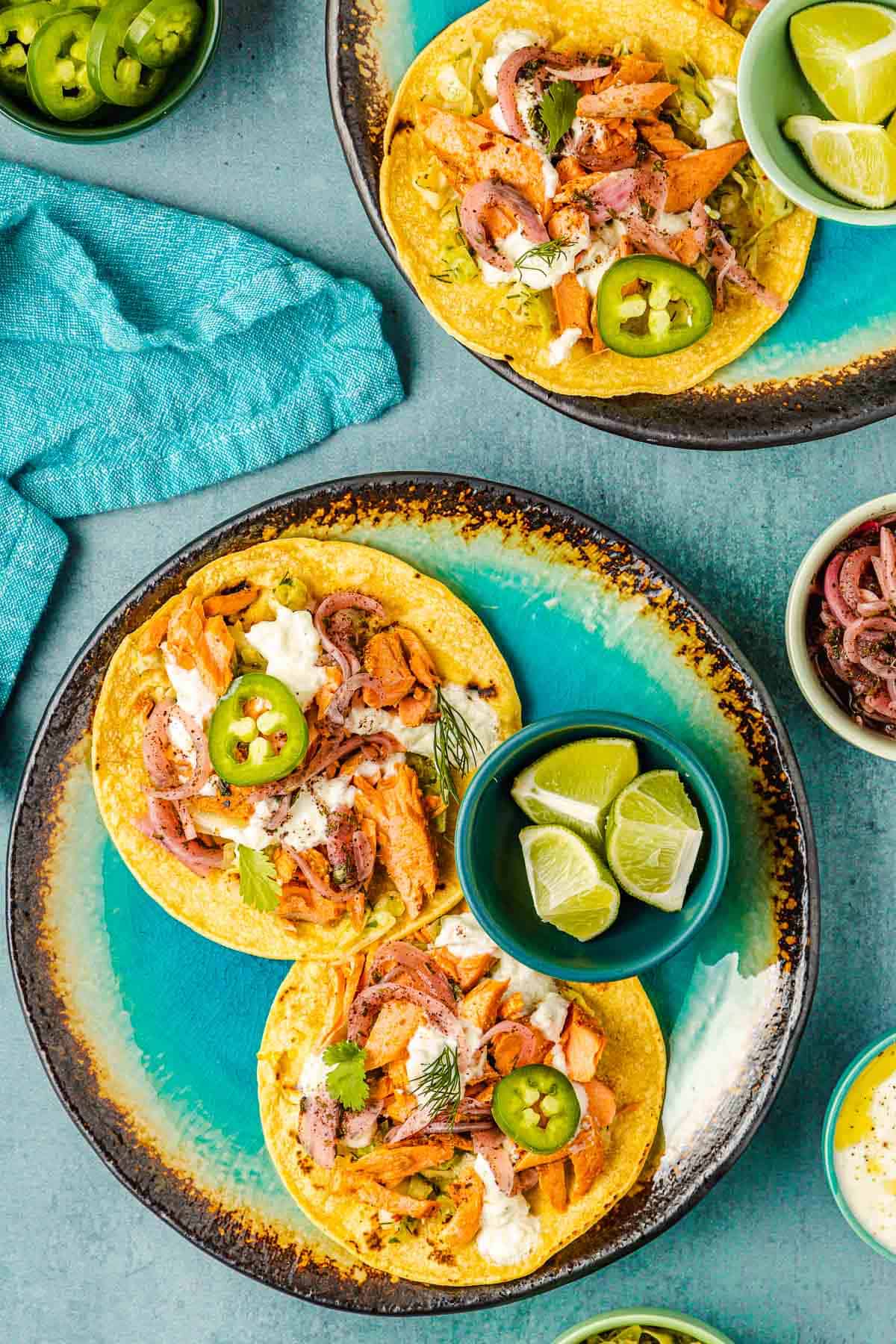 two open salmon tacos on corn tortillas and a bowl of 3 lime wedges on a plate, with another plate of salmon tacos and a bowl of sliced jalapenos in the background.