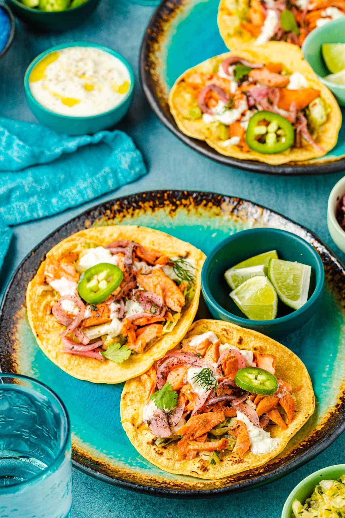 two open salmon tacos on corn tortillas and a bowl of 3 lime wedges on a plate, with another plate of salmon tacos and a bowl of tzatziki in the background.