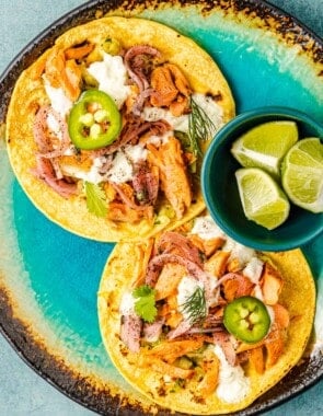 two open salmon tacos on corn tortillas topped with tzatziki, pickled onions and sliced jalapenos on a plate with a bowl of 3 lime wedges.