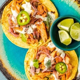 two open salmon tacos on corn tortillas topped with tzatziki, pickled onions and sliced jalapenos on a plate with a bowl of 3 lime wedges.