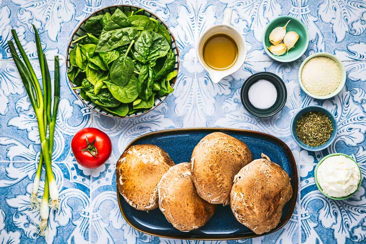 ingredients for stuffed portobello mushrooms including portobello mushrooms, olive oil, salt, italian seasoning, tomato, garlic, baby spinach, green onions ricotta cheese, and breadcrumbs.