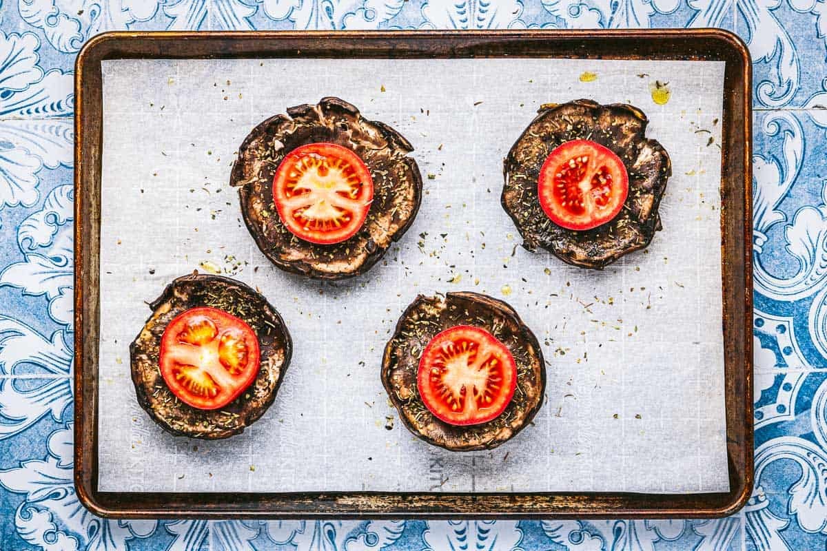 portobello mushroom caps topped with tomato slices on a parchment lined baking sheet.