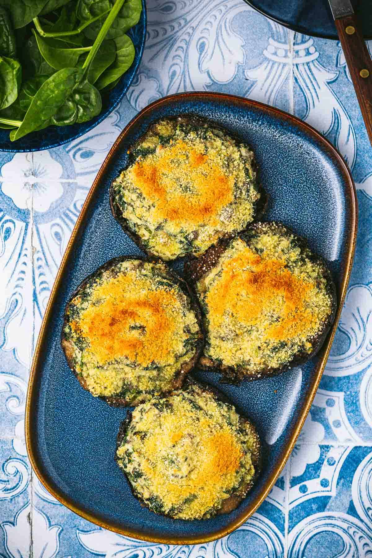 4 cooked stuffed portobello mushrooms on a serving platter next to a bowl of baby spinach.
