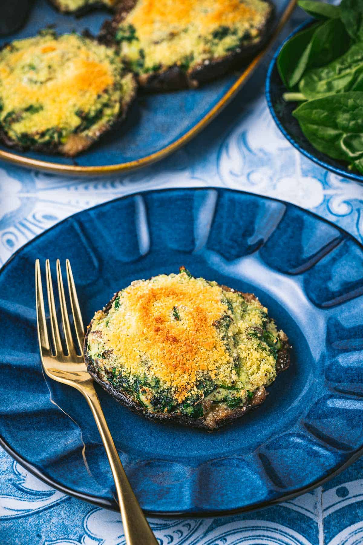 a cooked stuffed portobello mushroom on a plate with a fork next to a platter of stuffed portobello mushrooms and a bowl of baby spinach.