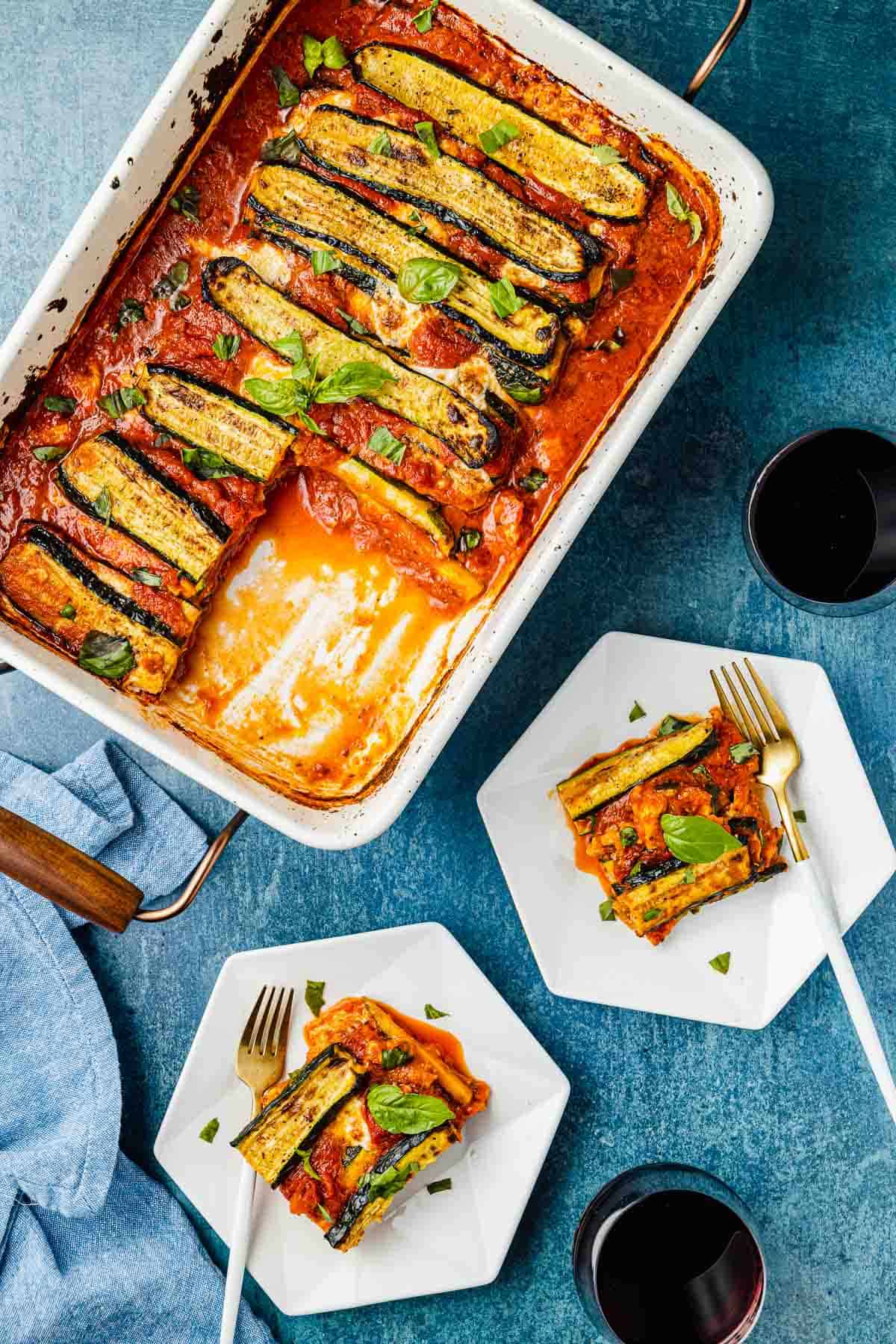 Baked zucchini parmesan in a baking dish next to two plates, each containing one slice of zucchini parmesan and a fork, and two glasses of red wine.