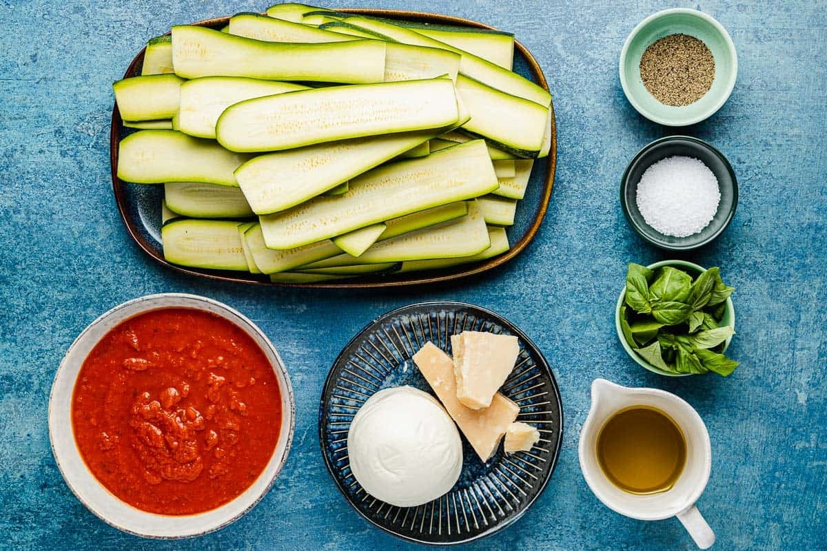 ingredients for zucchini parmesan including long slices of zucchini, salt, pepper, red pasta sauce, mozzarella, parmesan, olive oil and fresh basil leaves.