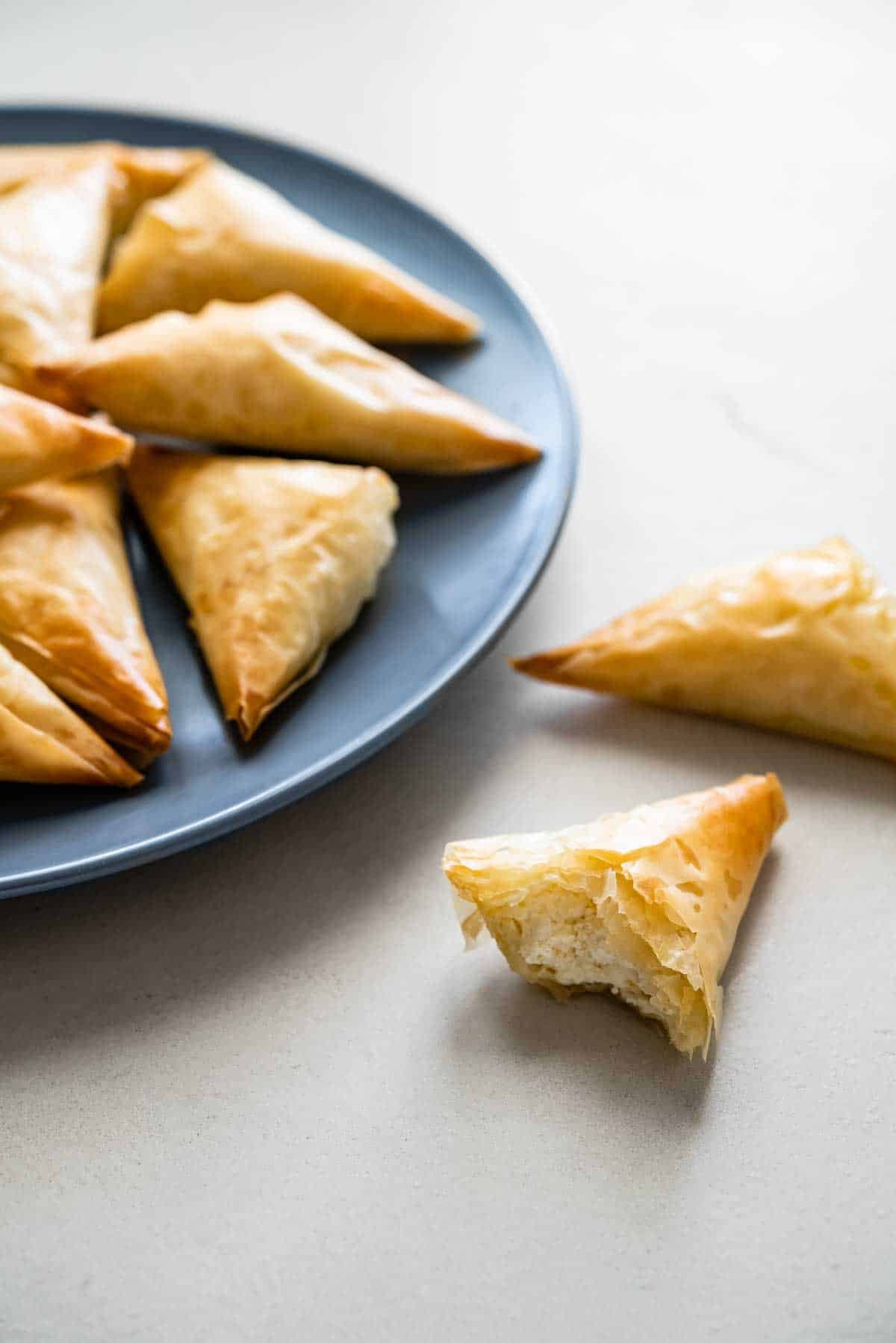 two tiropitakia feta hand pie halves next to a plate full of tiropitakia.