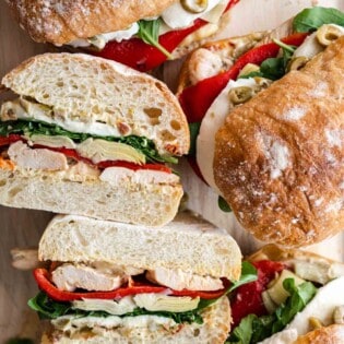 four antipasto chicken sandwiches on a serving board, one cut in halve showing it's ingredients.