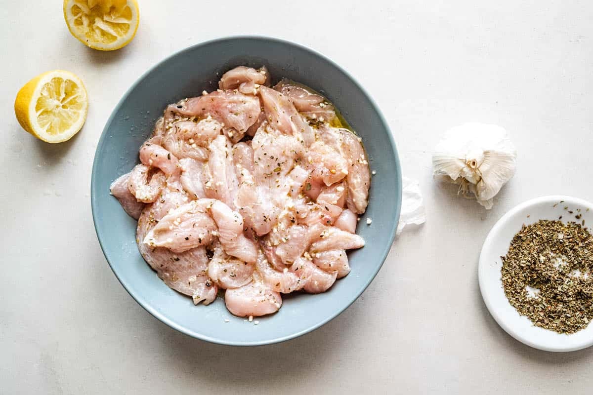 sliced raw chicken breast marinating in a bowl next to 2 lemon halves, a head of garlic and a bowl of oregano.