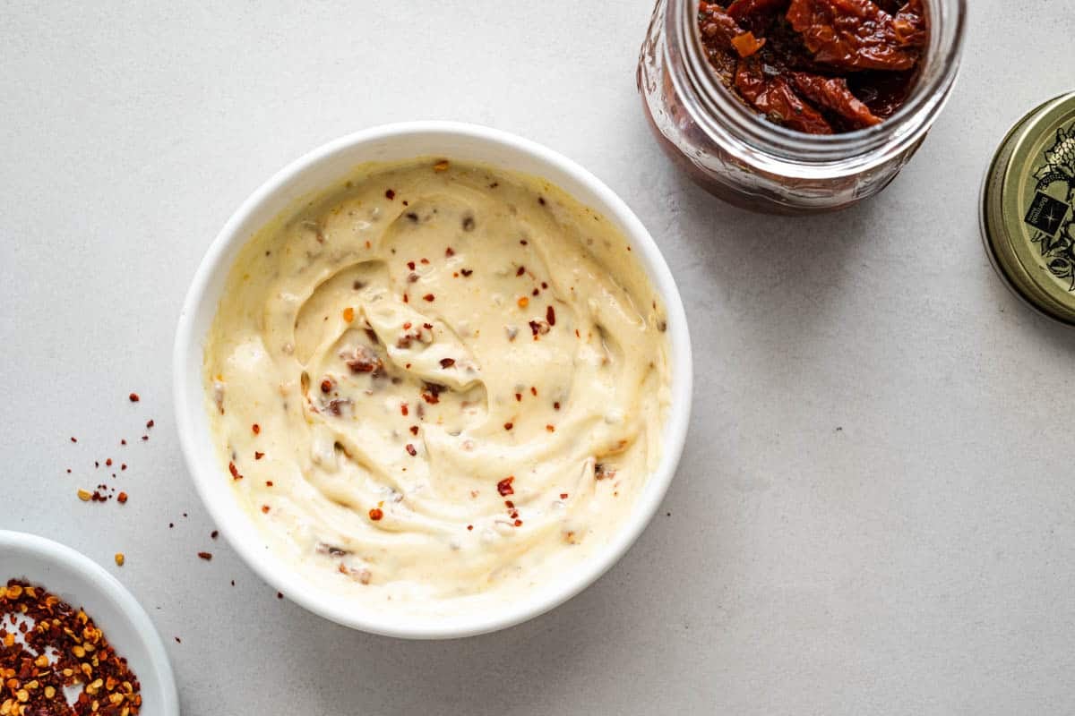 a bowl of sun-dried tomato aioli next to a jar of sun-dried tomatoes and a bowl of red pepper flakes.