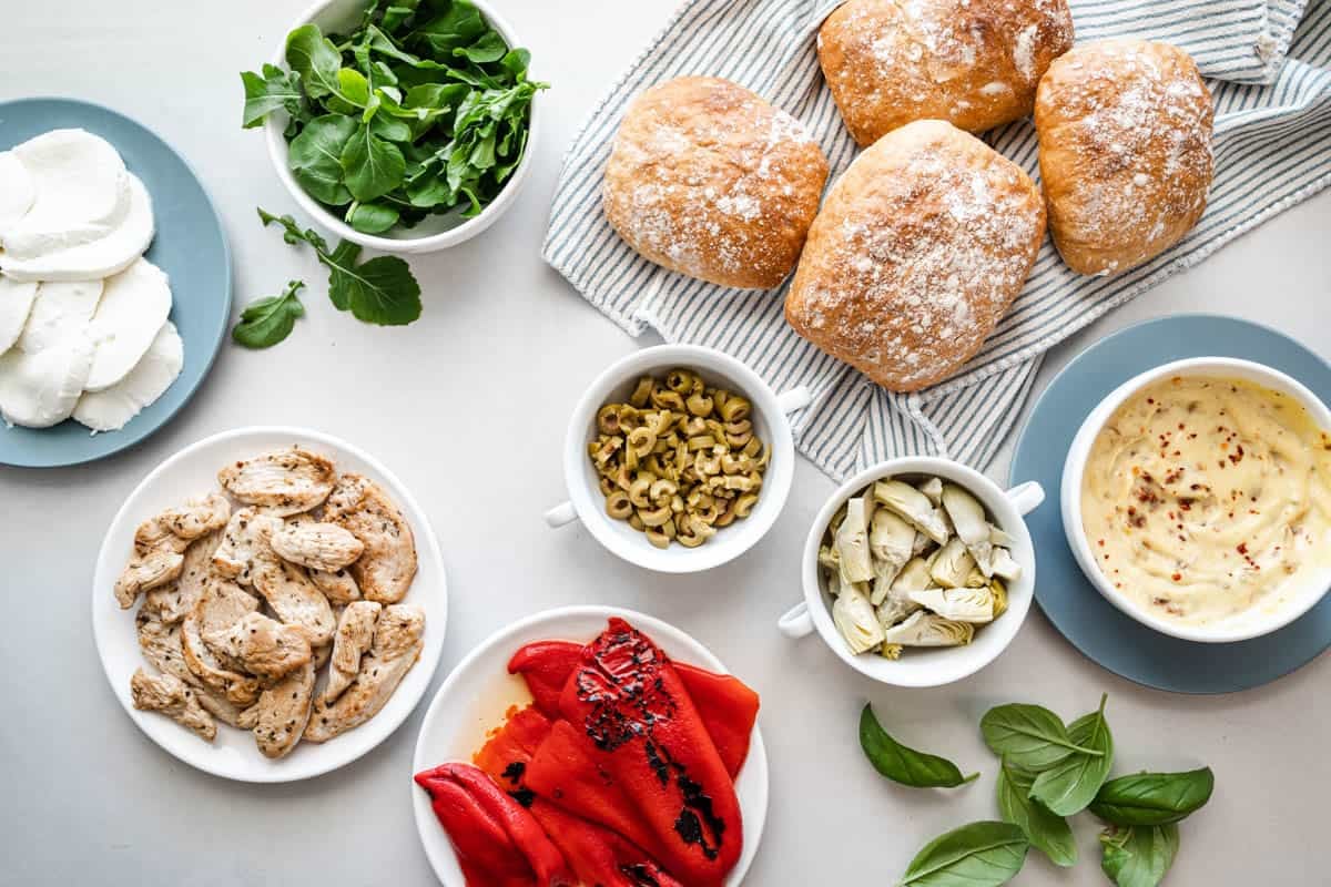 ingredients for an antipasto chicken sandwich including chicken breast, roasted red peppers, basil, sliced mozzarella, arugula, sliced green olives, artichoke hearts, ciabatta rolls and sundried tomato aioli.