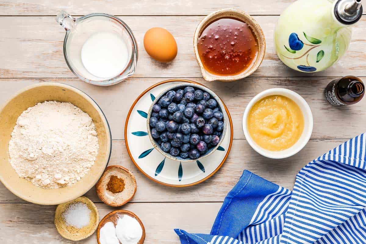 ingredients for healthy blueberry muffins including white whole wheat flour, baking powder, baking soda, salt, cinnamon, honey, apple sauce, olive oil, milk, an egg, vanilla extract and blueberries.