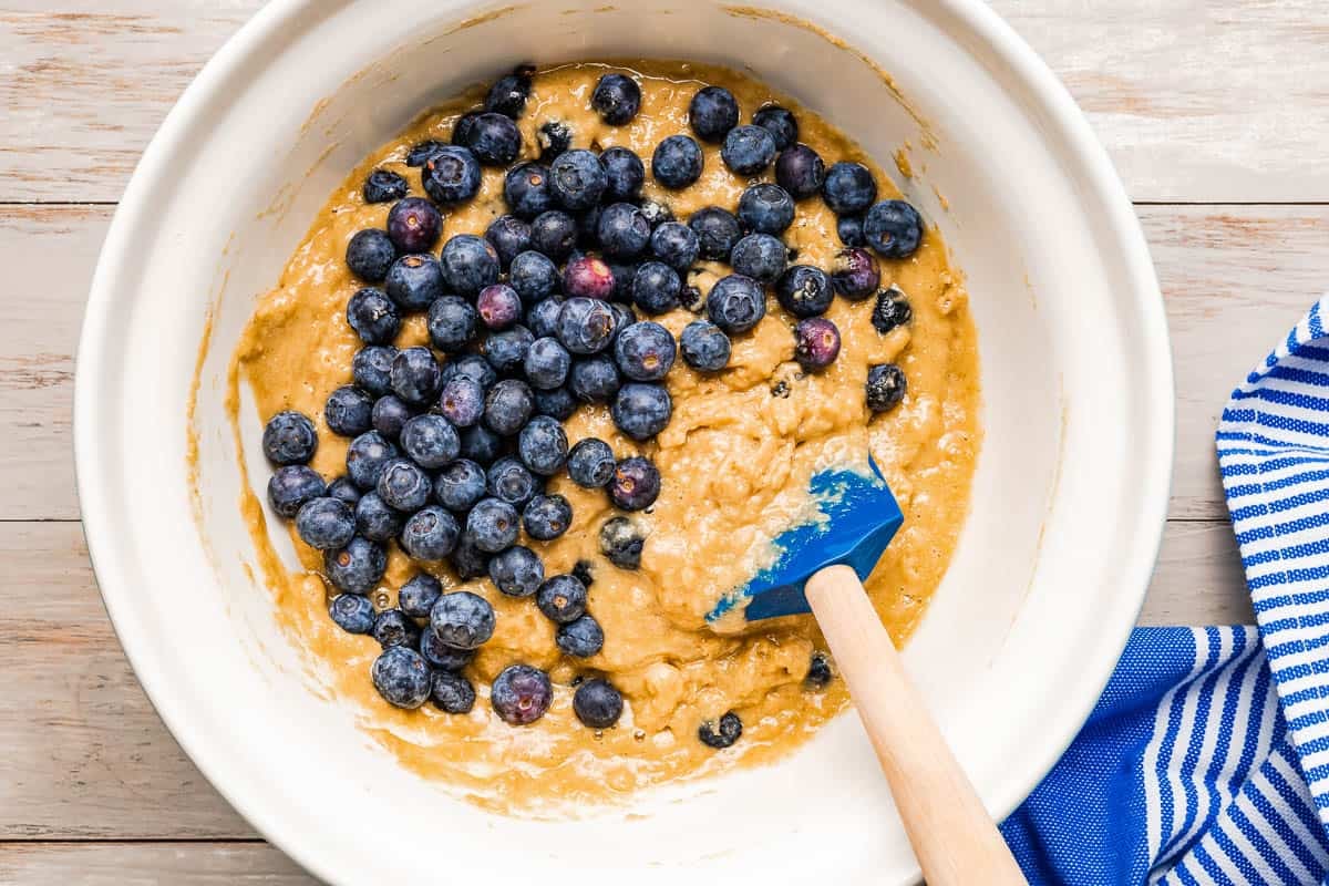 blueberries added to blueberry muffin batter in a bowl with a rubber spatula.