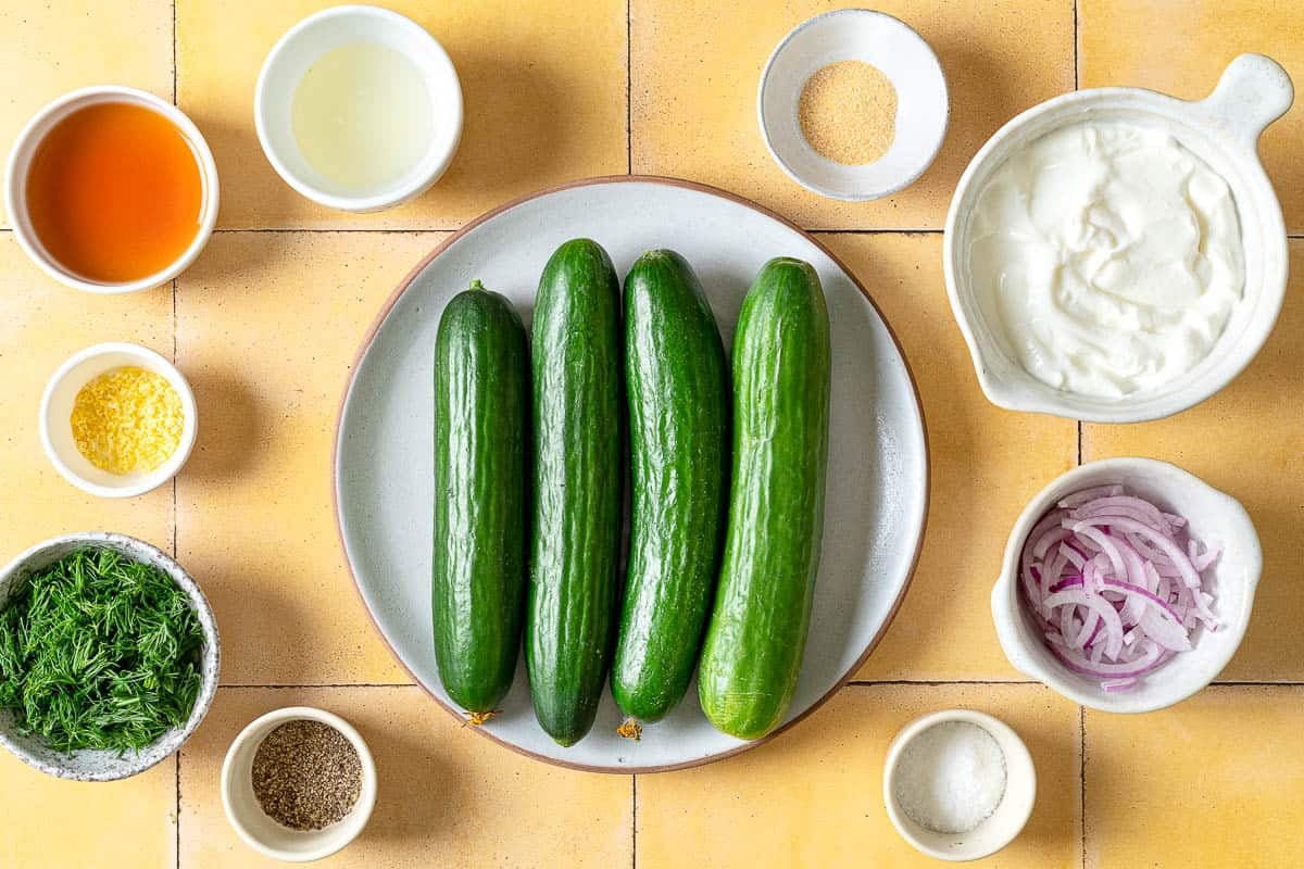 ingredients for creamy cucumber salad including cucumbers, salt, greek yogurt, red wine vinegar, lemon zest, lemon juice, fresh dill, garlic powder, pepper and sliced red onion.