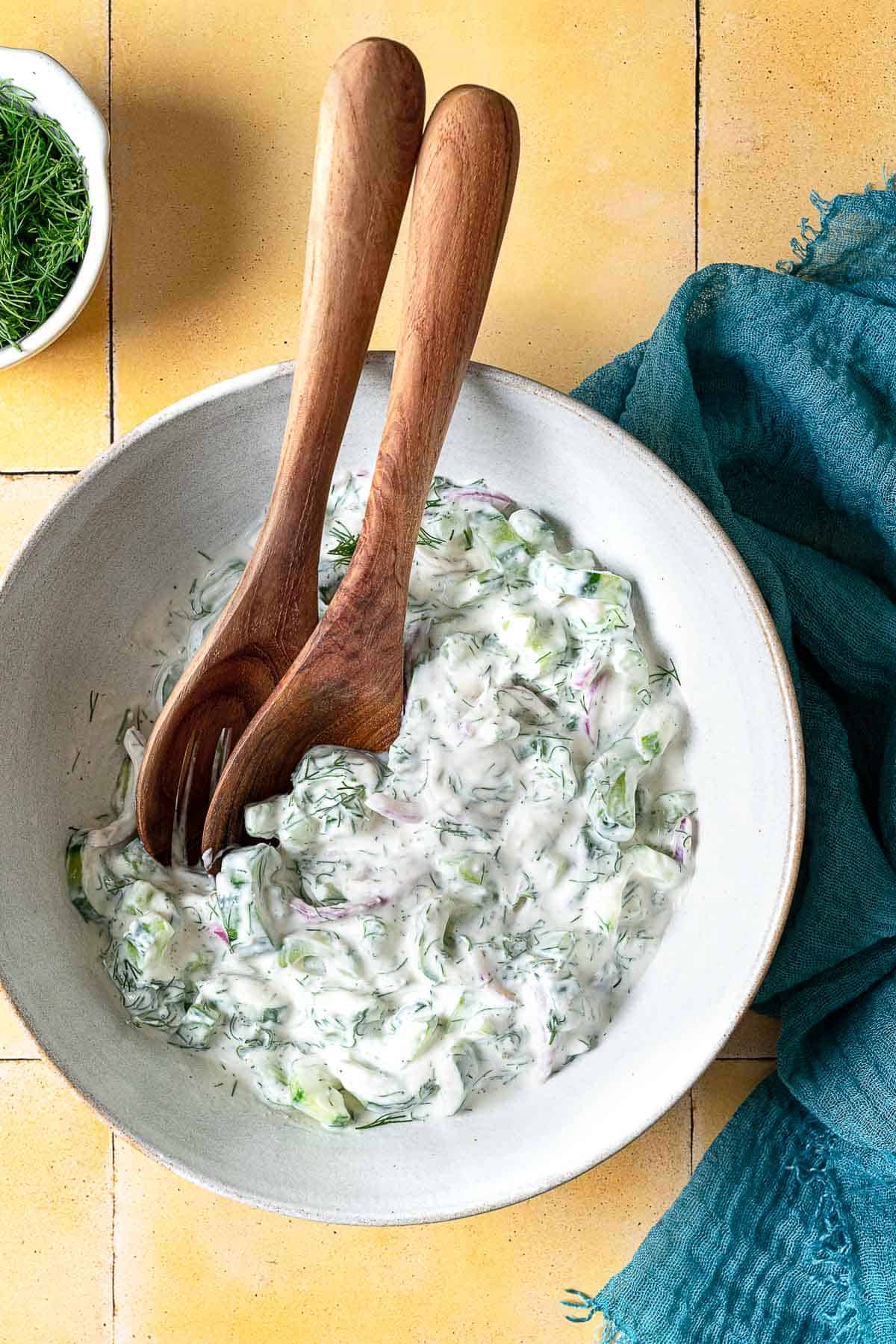 creamy cucumber salad in a serving bowl with wooden serving utensils next to a bowl of dill.
