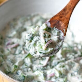 a bite of creamy cucumber salad being lifted out of a serving bowl with a wooden serving spoon.