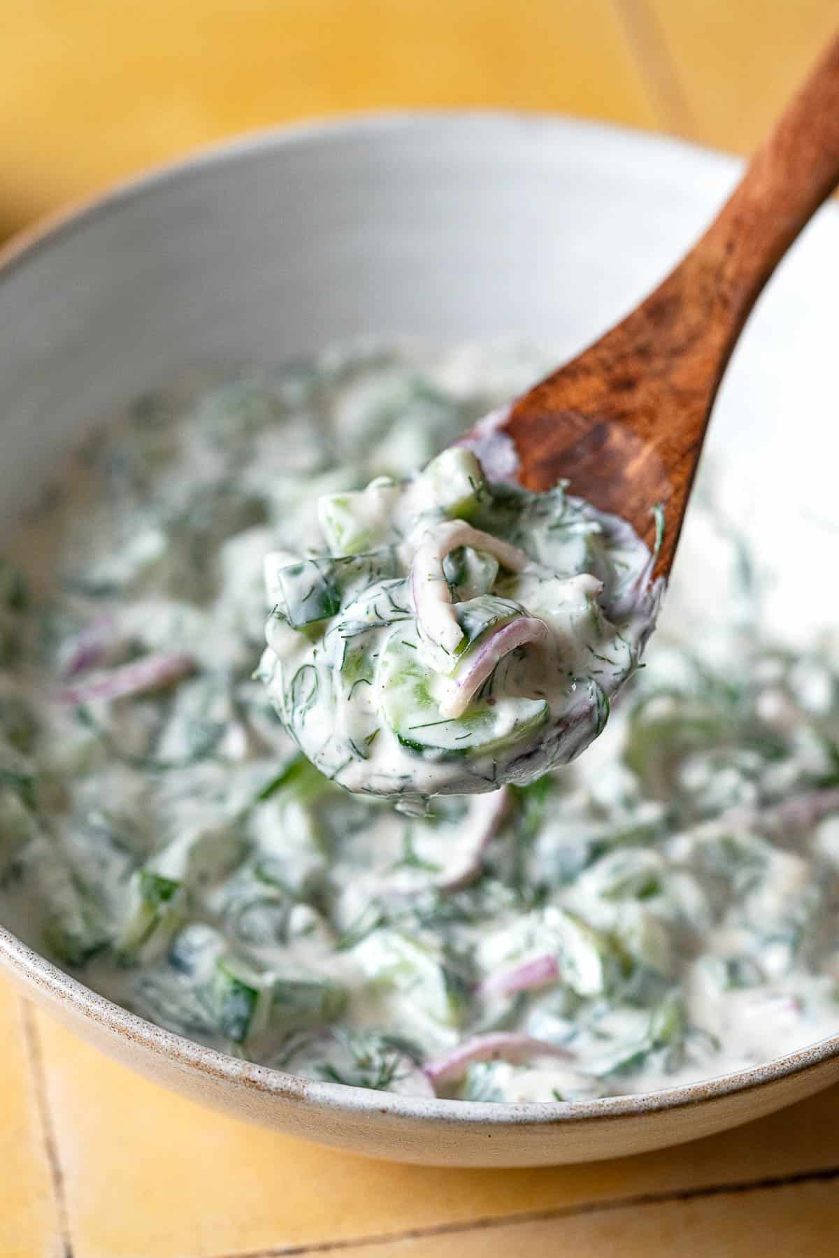 a bite of creamy cucumber salad being lifted out of a serving bowl with a wooden serving spoon.