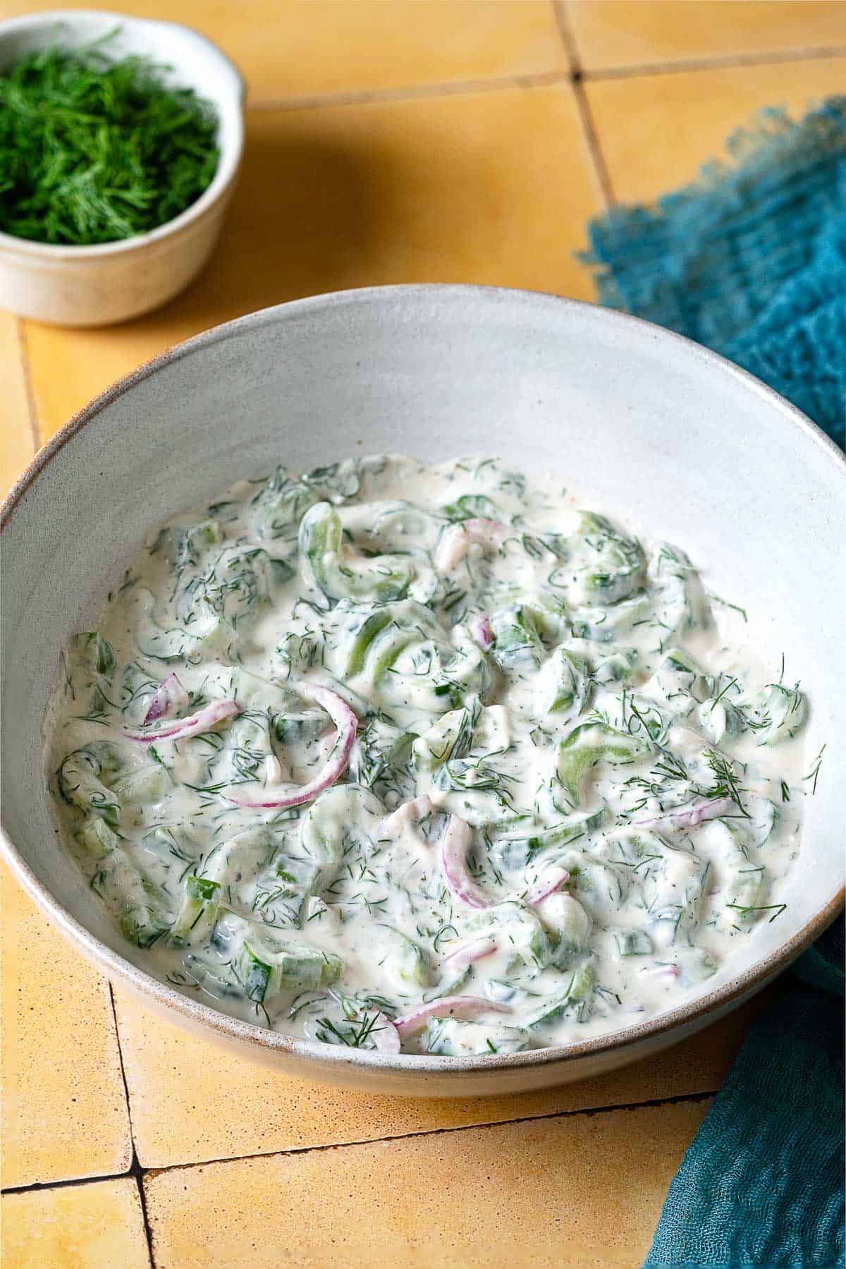 a creamy cucumber salad in a serving bowl next to a bowl of dill.