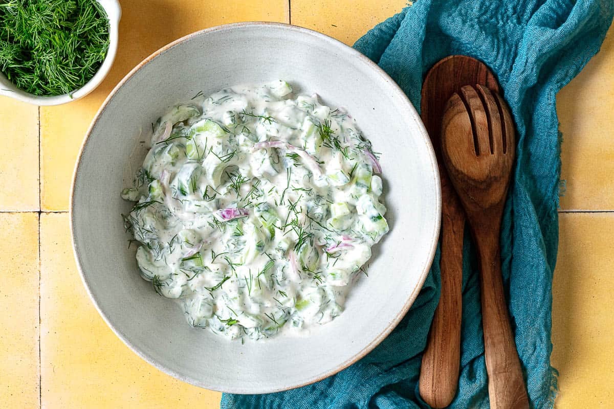 overhead photo of a creamy cucumber salad in a serving bowl next to wooden serving utensils and a bowl of dill.