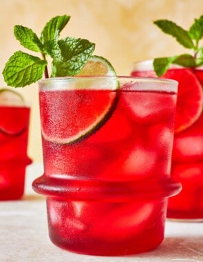 hibiscus iced tea in a glass garnished of mint and lime with two more glasses of hibiscus tea in the background.