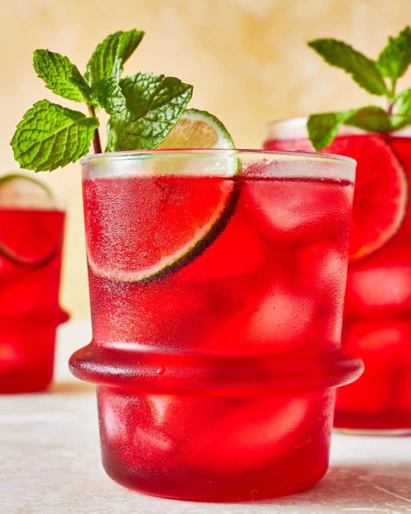 hibiscus iced tea in a glass garnished of mint and lime with two more glasses of hibiscus tea in the background.