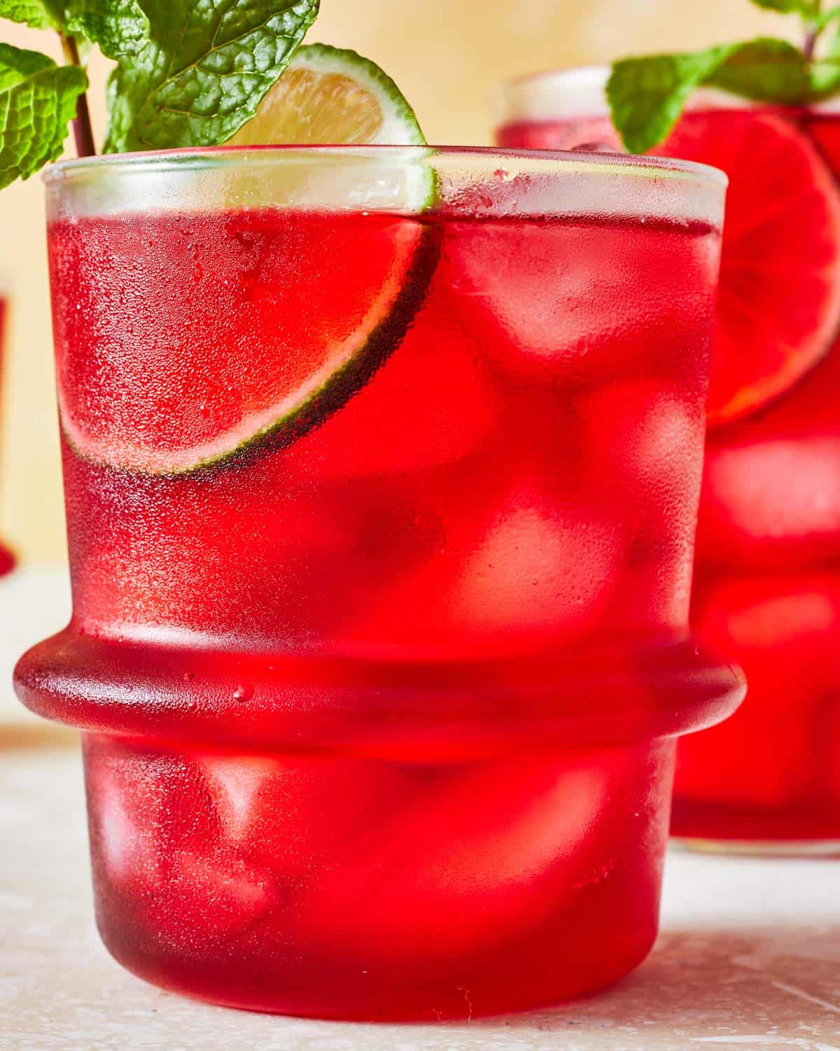 hibiscus iced tea in a glass garnished of mint and lime with another glass of hibiscus tea in the background.