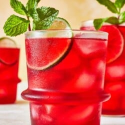 hibiscus iced tea in a glass garnished of mint and lime with two more glasses of hibiscus tea in the background.