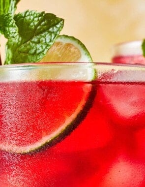 close up of hibiscus iced tea in a glass garnished of mint and lime with two more glasses of hibiscus tea in the background.