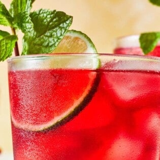 close up of hibiscus iced tea in a glass garnished of mint and lime with two more glasses of hibiscus tea in the background.