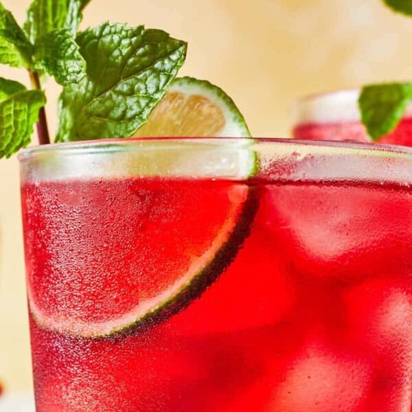 close up of hibiscus iced tea in a glass garnished of mint and lime with two more glasses of hibiscus tea in the background.